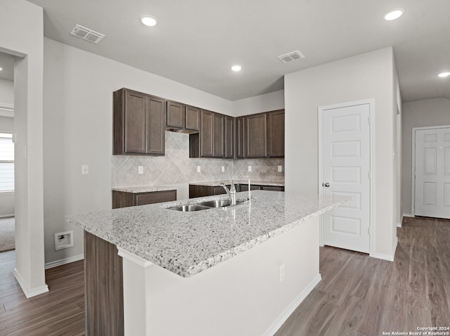 kitchen featuring wood-type flooring, light stone counters, a kitchen island with sink, sink, and decorative backsplash