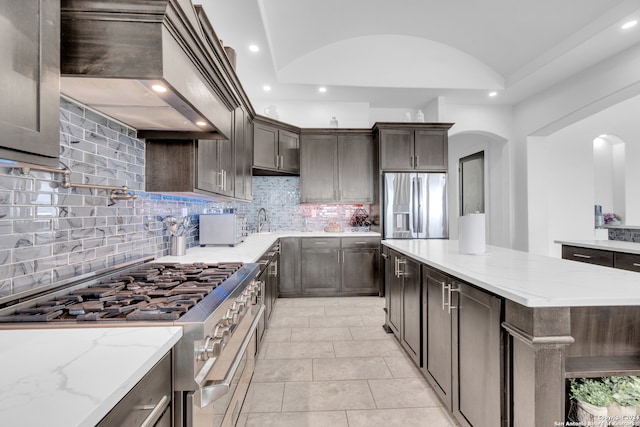 kitchen with tasteful backsplash, stainless steel appliances, custom range hood, light stone countertops, and vaulted ceiling