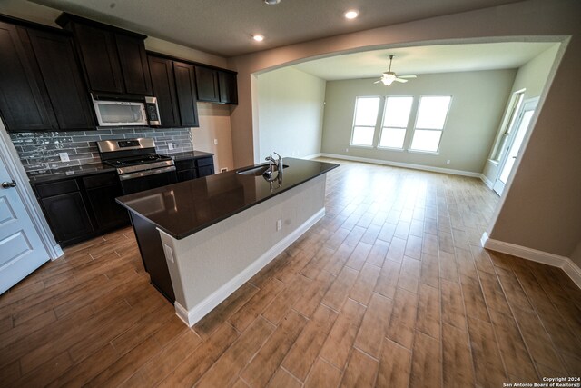 kitchen with light wood-type flooring, backsplash, a center island with sink, appliances with stainless steel finishes, and ceiling fan