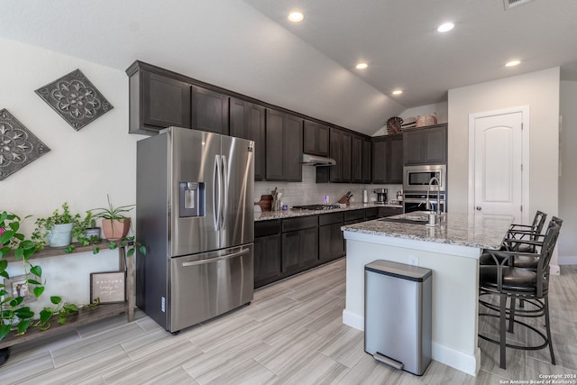 kitchen featuring appliances with stainless steel finishes, vaulted ceiling, light stone countertops, dark brown cabinets, and a kitchen island with sink