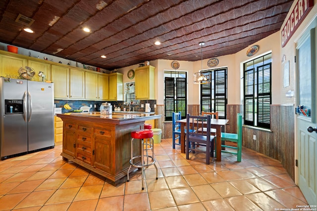 kitchen with light tile patterned floors, decorative light fixtures, stainless steel refrigerator with ice dispenser, light brown cabinetry, and a kitchen bar