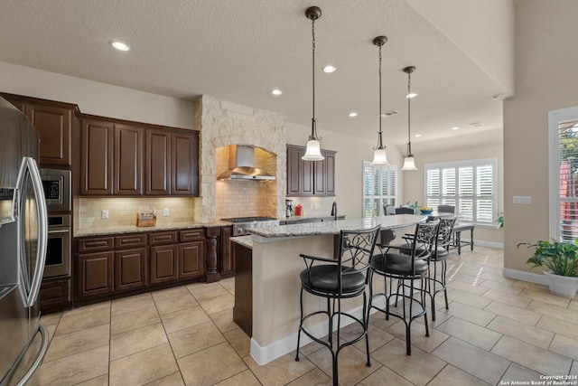 kitchen featuring pendant lighting, an island with sink, a textured ceiling, wall chimney range hood, and appliances with stainless steel finishes