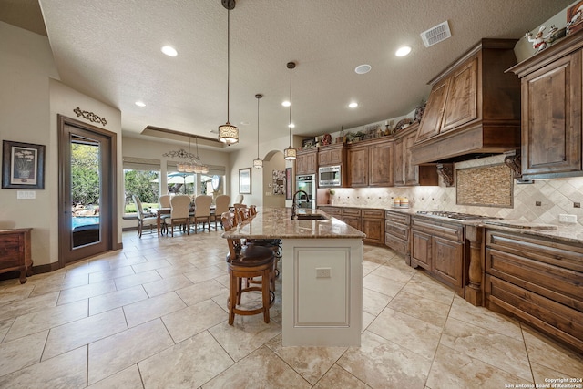 kitchen featuring pendant lighting, light stone counters, a textured ceiling, tasteful backsplash, and an island with sink