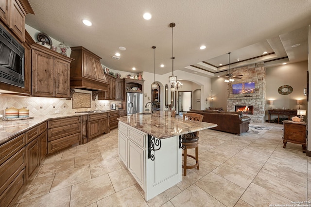 kitchen featuring custom exhaust hood, a raised ceiling, appliances with stainless steel finishes, a spacious island, and a fireplace