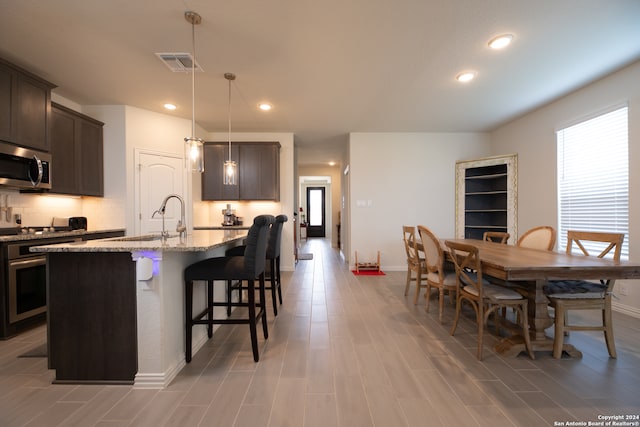 kitchen featuring a kitchen island with sink, sink, stainless steel appliances, decorative light fixtures, and light stone countertops