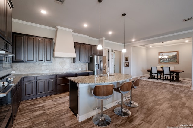 kitchen with wood-type flooring, premium range hood, a center island with sink, and stainless steel fridge