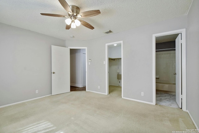 unfurnished bedroom with a spacious closet, a textured ceiling, ceiling fan, ensuite bath, and light carpet