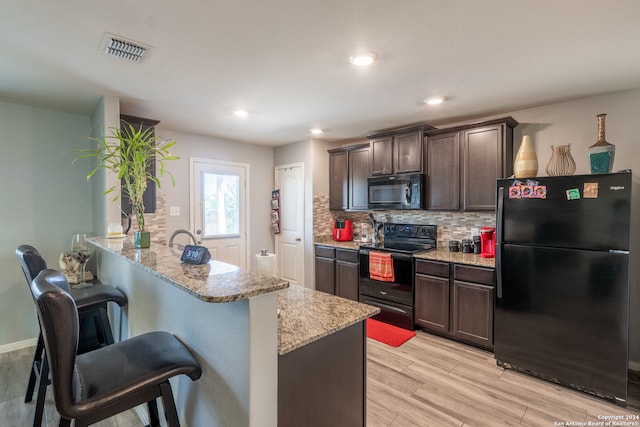 kitchen featuring light wood-type flooring, dark brown cabinets, black appliances, a breakfast bar area, and light stone countertops