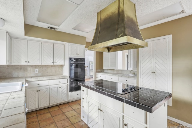 kitchen with tasteful backsplash, island range hood, black appliances, white cabinetry, and tile countertops