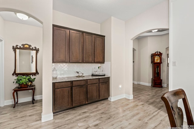 bar with dark brown cabinets, light wood-type flooring, decorative backsplash, and light stone countertops