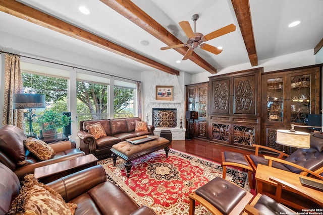 living room featuring wood-type flooring, beam ceiling, ceiling fan, and a stone fireplace