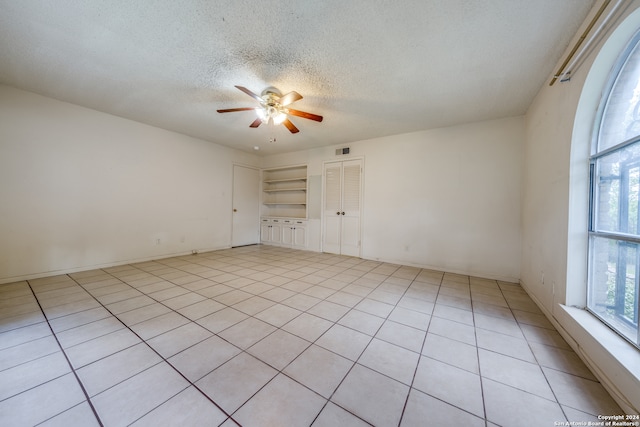 tiled empty room featuring a textured ceiling, built in shelves, and ceiling fan