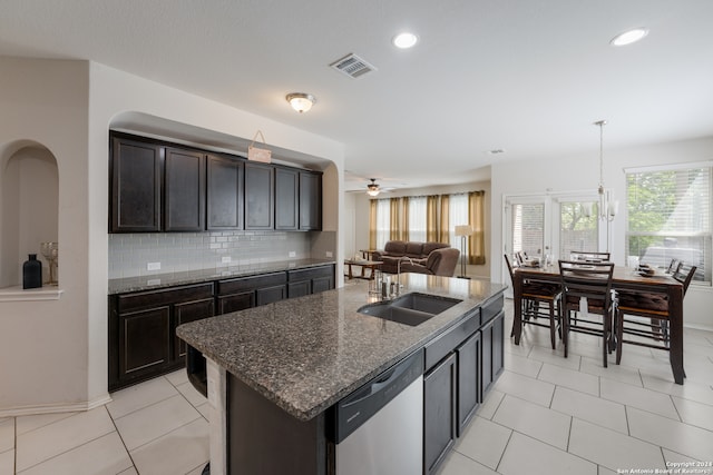 kitchen featuring dark stone countertops, tasteful backsplash, ceiling fan with notable chandelier, stainless steel dishwasher, and a kitchen island with sink