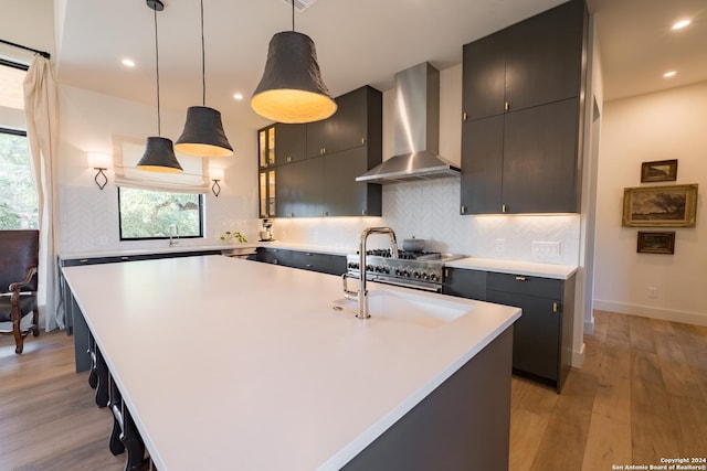 kitchen with backsplash, wall chimney range hood, light hardwood / wood-style flooring, an island with sink, and decorative light fixtures