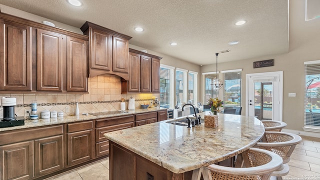 kitchen with hanging light fixtures, a textured ceiling, a kitchen island with sink, light stone countertops, and a kitchen bar