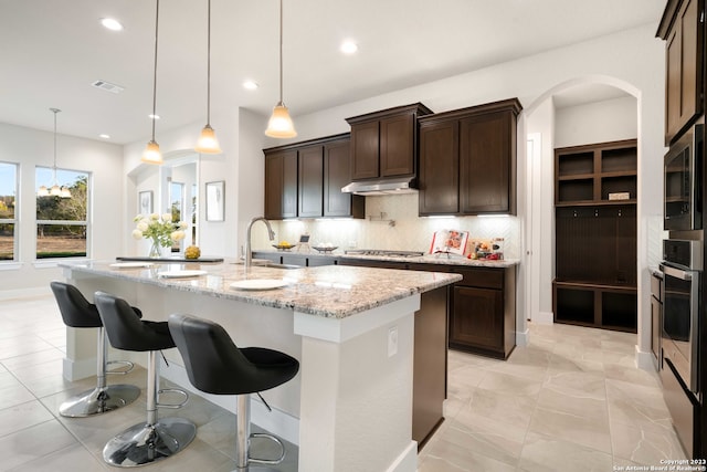 kitchen featuring an island with sink, hanging light fixtures, stainless steel appliances, light stone countertops, and decorative backsplash