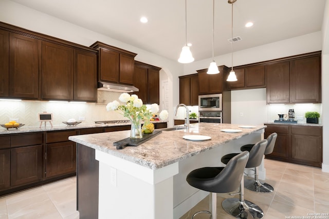 kitchen with appliances with stainless steel finishes, light stone counters, hanging light fixtures, and a kitchen island with sink