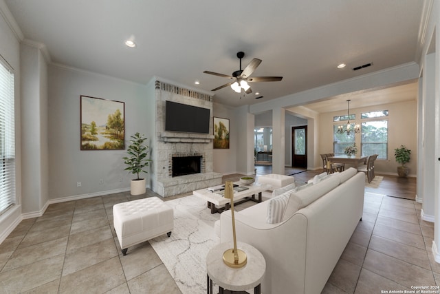 tiled living room with ceiling fan with notable chandelier, ornamental molding, and a stone fireplace