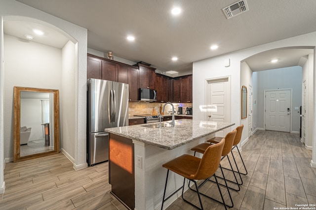 kitchen with light wood-type flooring, a kitchen island with sink, sink, stainless steel appliances, and light stone countertops