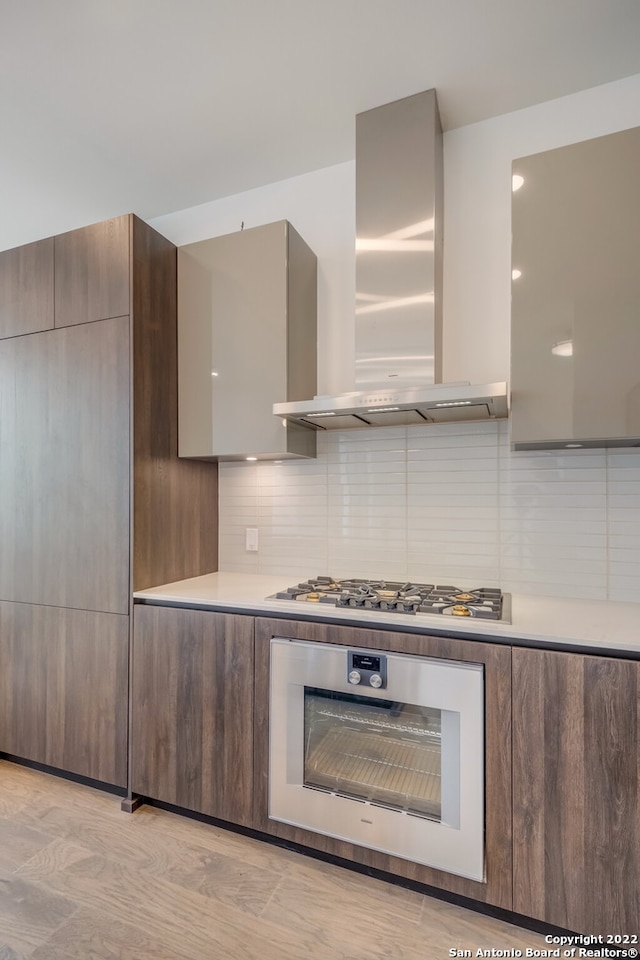 kitchen with light wood-type flooring, decorative backsplash, wall chimney exhaust hood, and stainless steel appliances