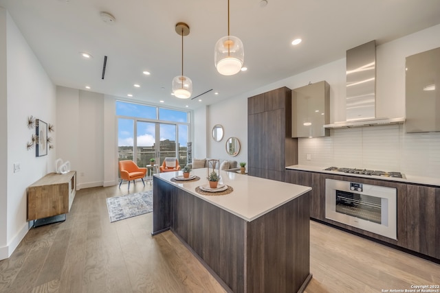 kitchen with light hardwood / wood-style floors, stainless steel appliances, a center island, decorative light fixtures, and wall chimney range hood