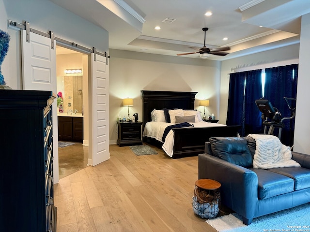 bedroom featuring a barn door, light wood-type flooring, a tray ceiling, ceiling fan, and ensuite bathroom