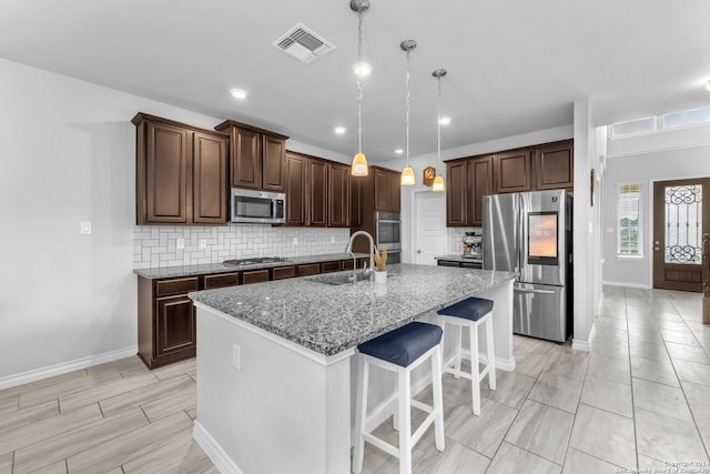 kitchen featuring light stone counters, stainless steel appliances, sink, hanging light fixtures, and an island with sink