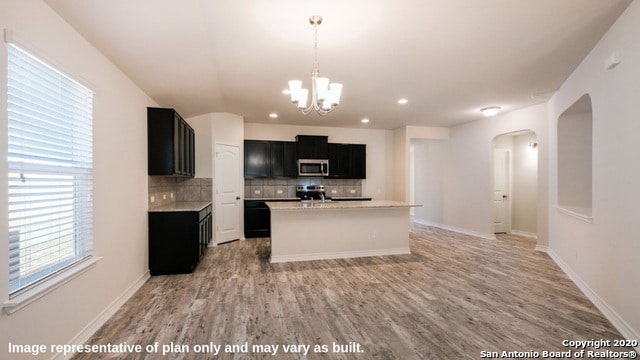 kitchen with appliances with stainless steel finishes, decorative backsplash, light wood-type flooring, decorative light fixtures, and a center island with sink