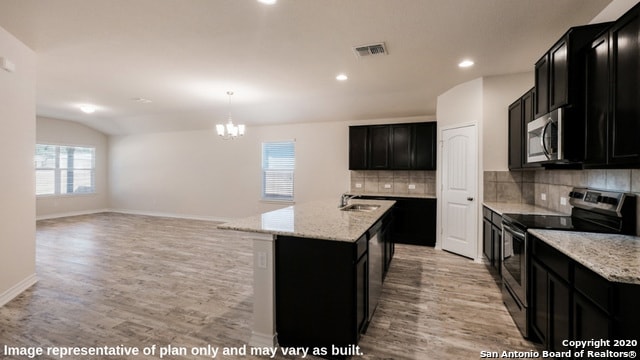 kitchen with pendant lighting, light stone countertops, a kitchen island with sink, appliances with stainless steel finishes, and light wood-type flooring