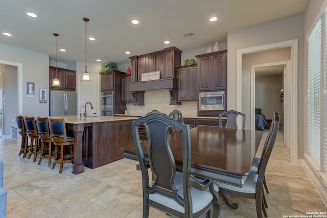 kitchen featuring appliances with stainless steel finishes, hanging light fixtures, decorative backsplash, dark brown cabinets, and a center island with sink