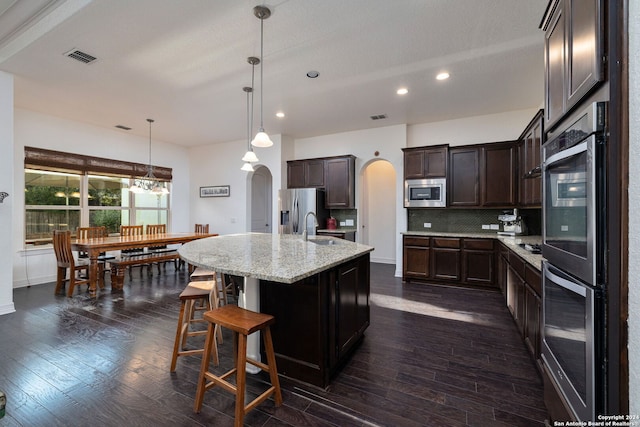 kitchen with light stone counters, pendant lighting, a kitchen island with sink, stainless steel appliances, and dark hardwood / wood-style flooring