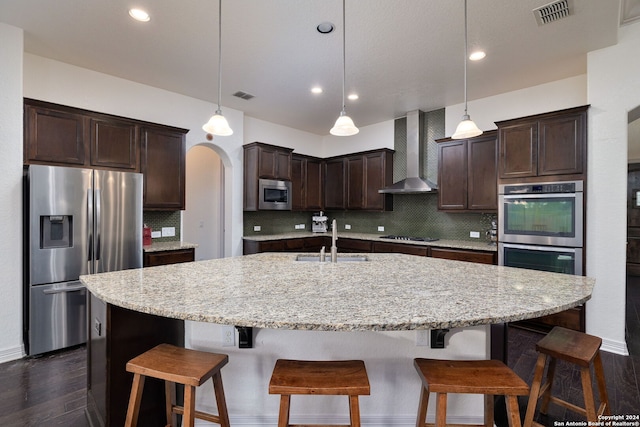 kitchen featuring sink, wall chimney exhaust hood, backsplash, a center island with sink, and appliances with stainless steel finishes