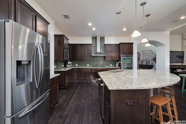 kitchen with dark wood-type flooring, tasteful backsplash, wall chimney exhaust hood, a center island with sink, and appliances with stainless steel finishes