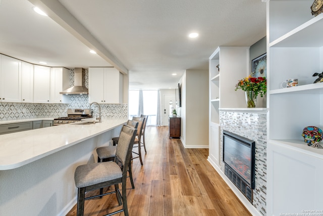 kitchen featuring a tile fireplace, stainless steel gas stove, wall chimney range hood, a kitchen bar, and white cabinets