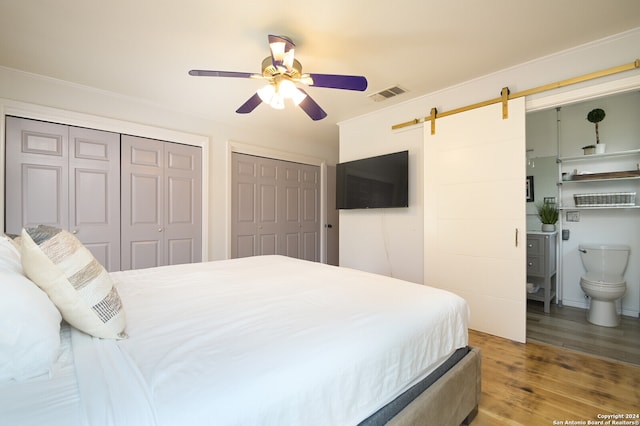 bedroom featuring a barn door, ensuite bathroom, hardwood / wood-style floors, two closets, and ceiling fan