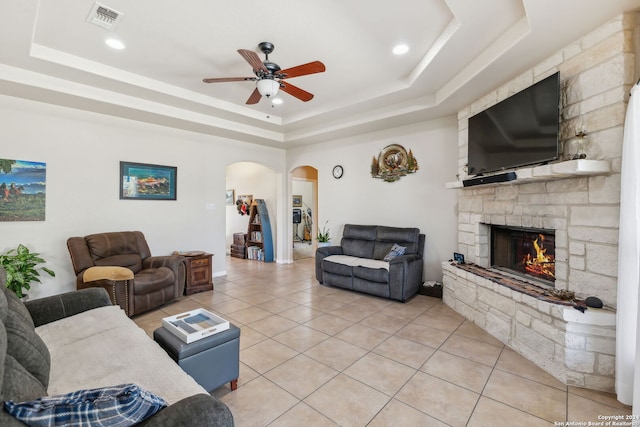 living room featuring a stone fireplace, a tray ceiling, ceiling fan, and tile patterned floors