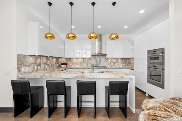 kitchen featuring dark wood-type flooring, white cabinets, kitchen peninsula, wall chimney exhaust hood, and decorative light fixtures