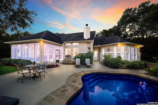 back of house at dusk with a patio area, a chimney, stucco siding, a sunroom, and an outdoor pool