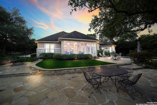 back of house at dusk featuring stucco siding, a patio, stone siding, outdoor dining area, and a yard