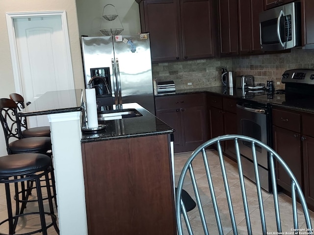 kitchen featuring dark brown cabinetry, a kitchen island with sink, stainless steel appliances, dark stone counters, and decorative backsplash