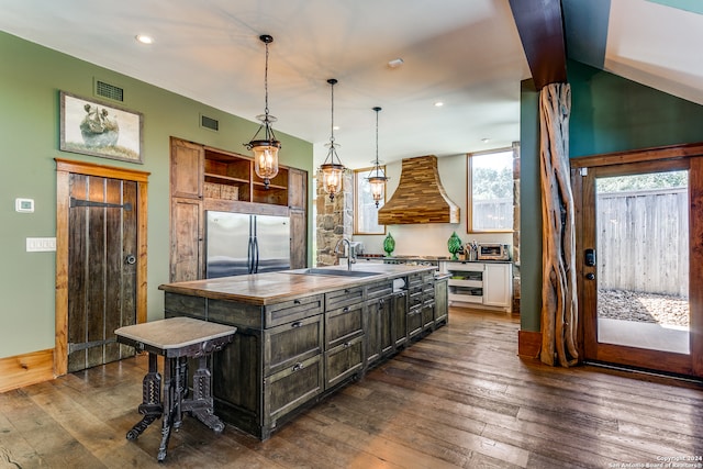 kitchen featuring pendant lighting, a center island with sink, built in fridge, dark hardwood / wood-style floors, and custom range hood