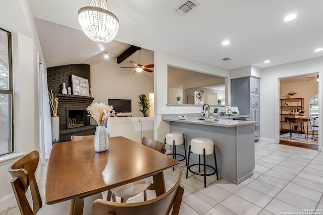 dining space featuring a fireplace, ceiling fan with notable chandelier, lofted ceiling with beams, and light tile patterned flooring