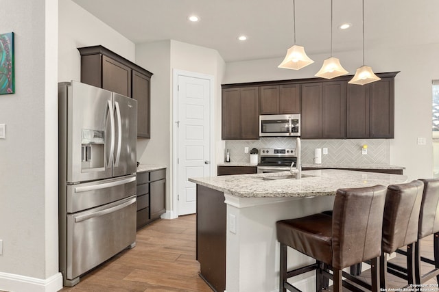 kitchen featuring appliances with stainless steel finishes, light stone counters, light hardwood / wood-style flooring, decorative light fixtures, and a kitchen island with sink