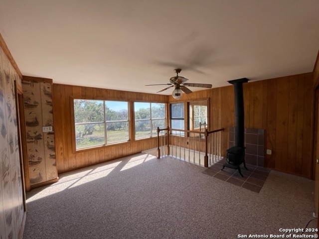 unfurnished living room featuring carpet flooring, wooden walls, ceiling fan, and a wood stove