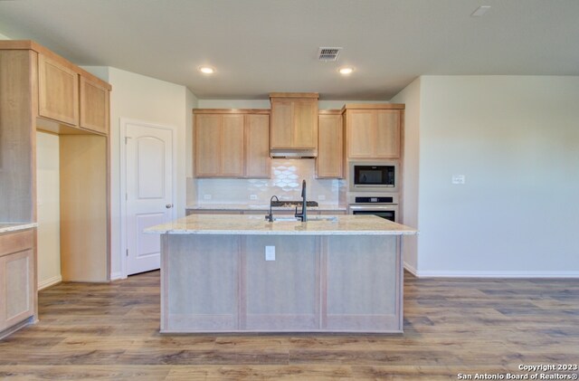 kitchen featuring light brown cabinets, stainless steel oven, built in microwave, and a kitchen island with sink