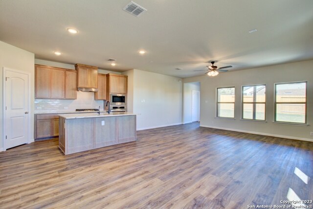 kitchen featuring built in microwave, light brown cabinets, backsplash, wood-type flooring, and a center island with sink