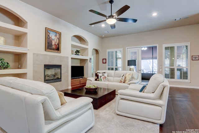 living room featuring wood-type flooring, a tile fireplace, built in shelves, and ceiling fan