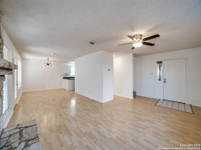 unfurnished living room with a textured ceiling, ceiling fan with notable chandelier, light hardwood / wood-style floors, and a stone fireplace