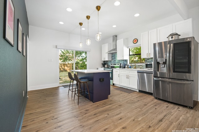 kitchen featuring white cabinets, wall chimney exhaust hood, stainless steel appliances, a center island, and decorative light fixtures