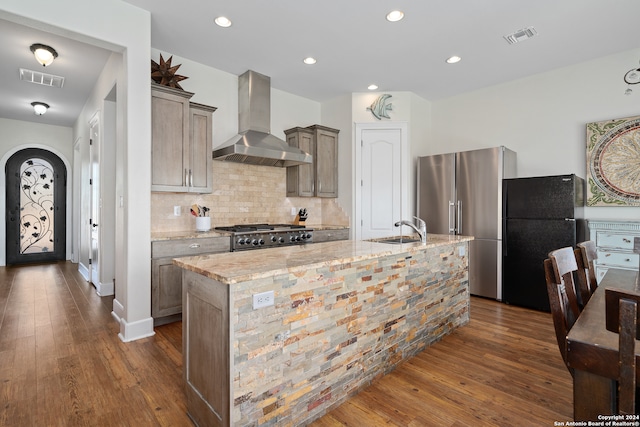 kitchen with light stone counters, wall chimney exhaust hood, a kitchen island with sink, appliances with stainless steel finishes, and dark hardwood / wood-style floors